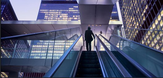 Person ascending an escalator between office buildings.